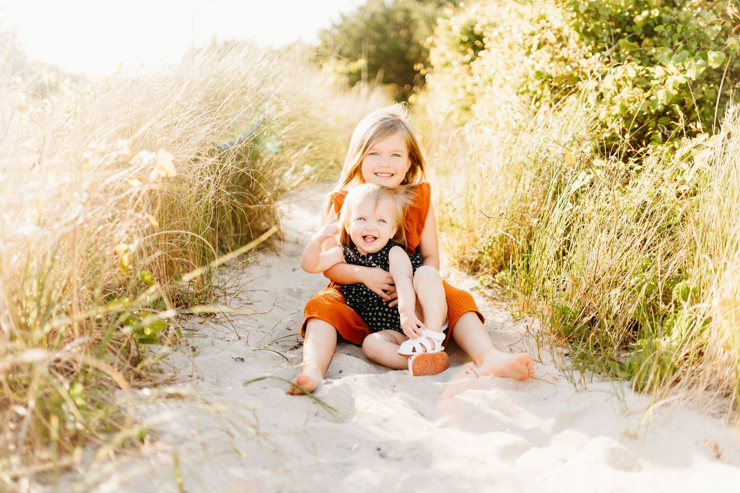 a big sister and little sister sitting in the sand smiling