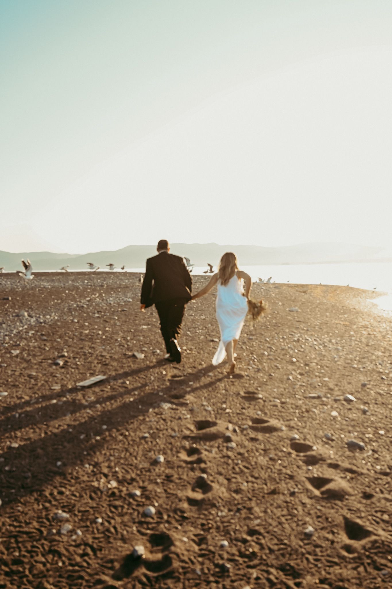 A couple strolls on a beach path at sunset casting long shadows on the ground.