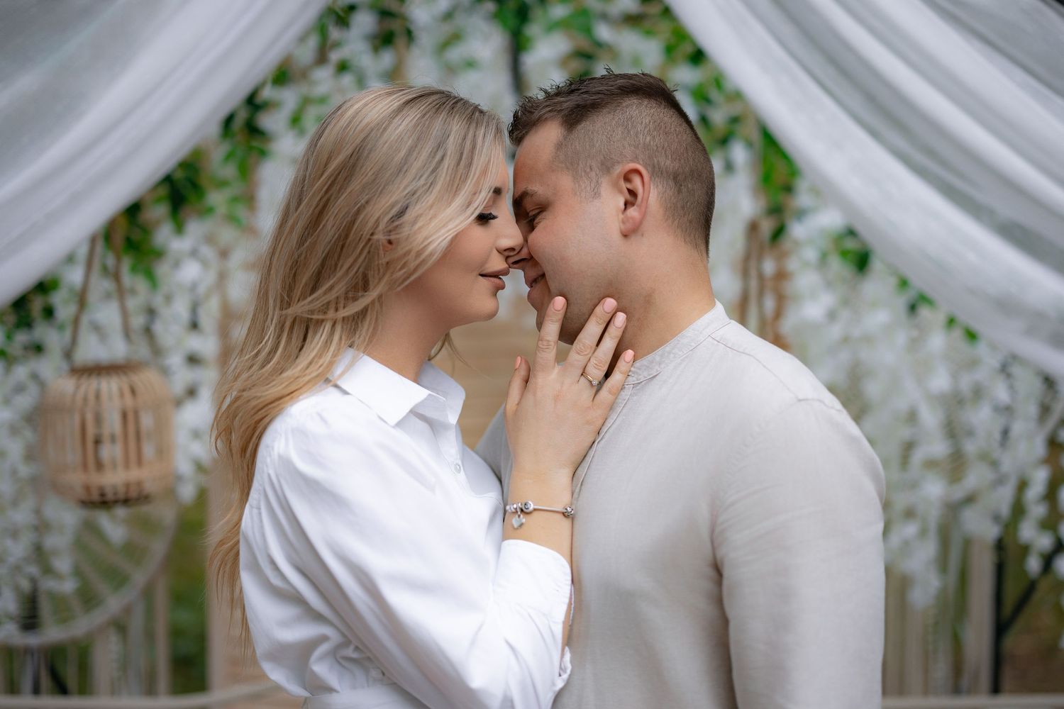 Couple in white outfits embracing under decorated wedding arch with hanging lanterns.