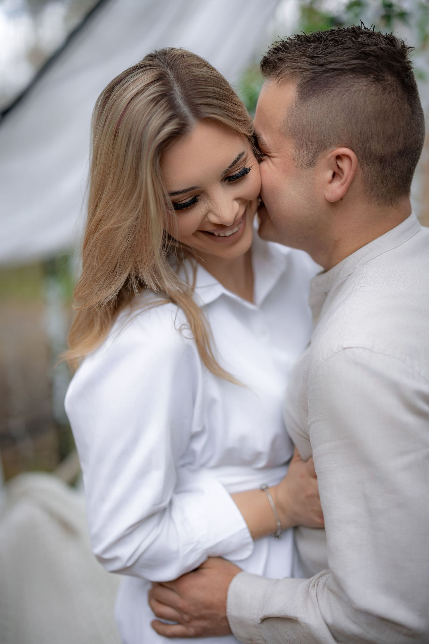 Intimate couple sharing tender moment in white decorated pergola with soft lighting.