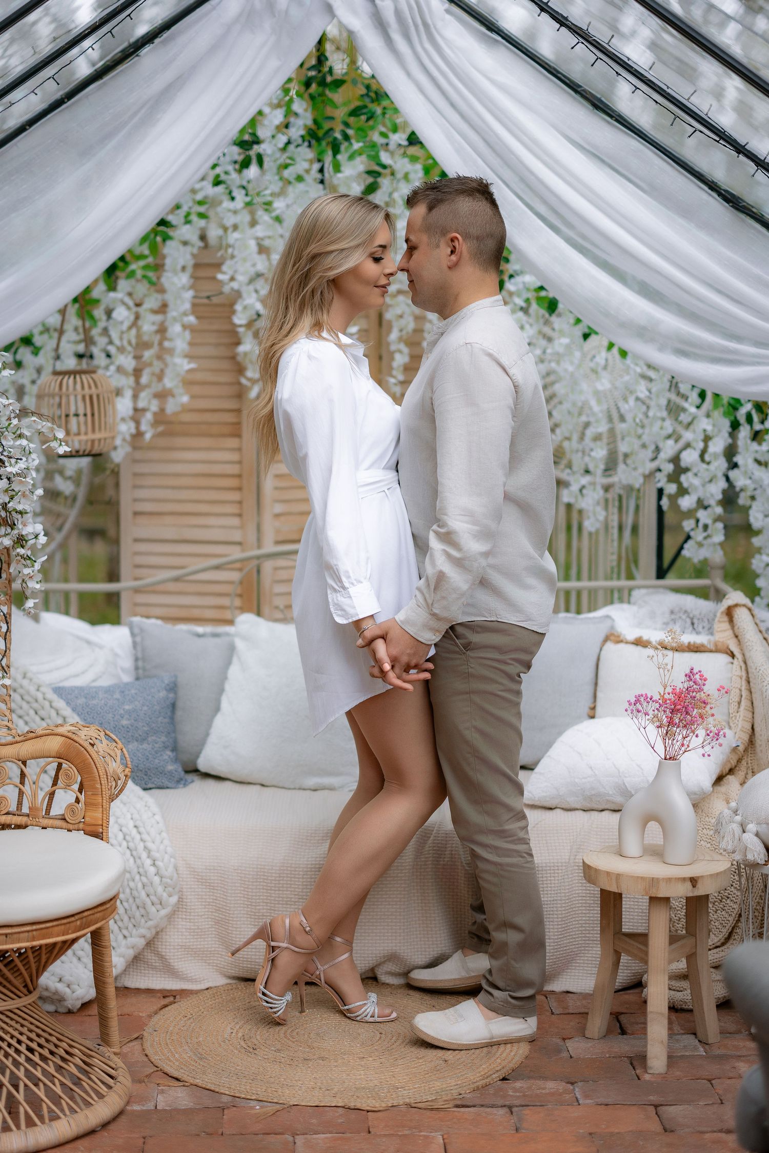 Couple in coordinating white outfits standing close together under draped canopy with greenery.