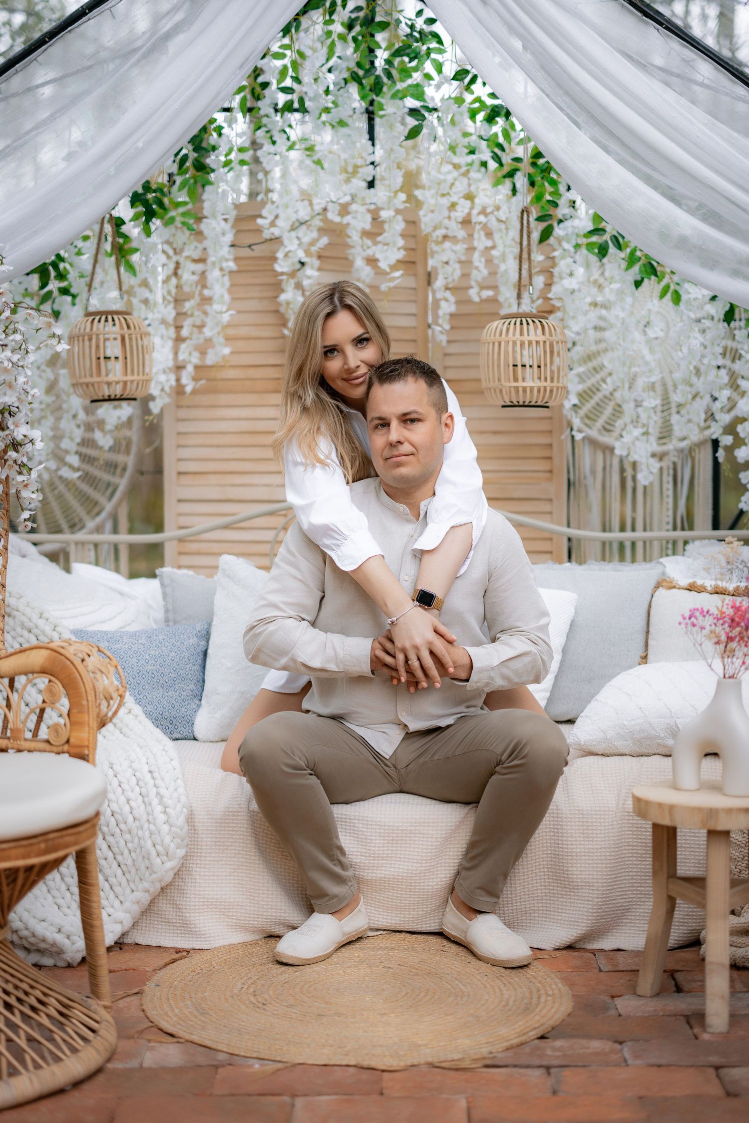 Couple relaxing on white sofa in garden gazebo decorated with hanging flowers and wicker accents.