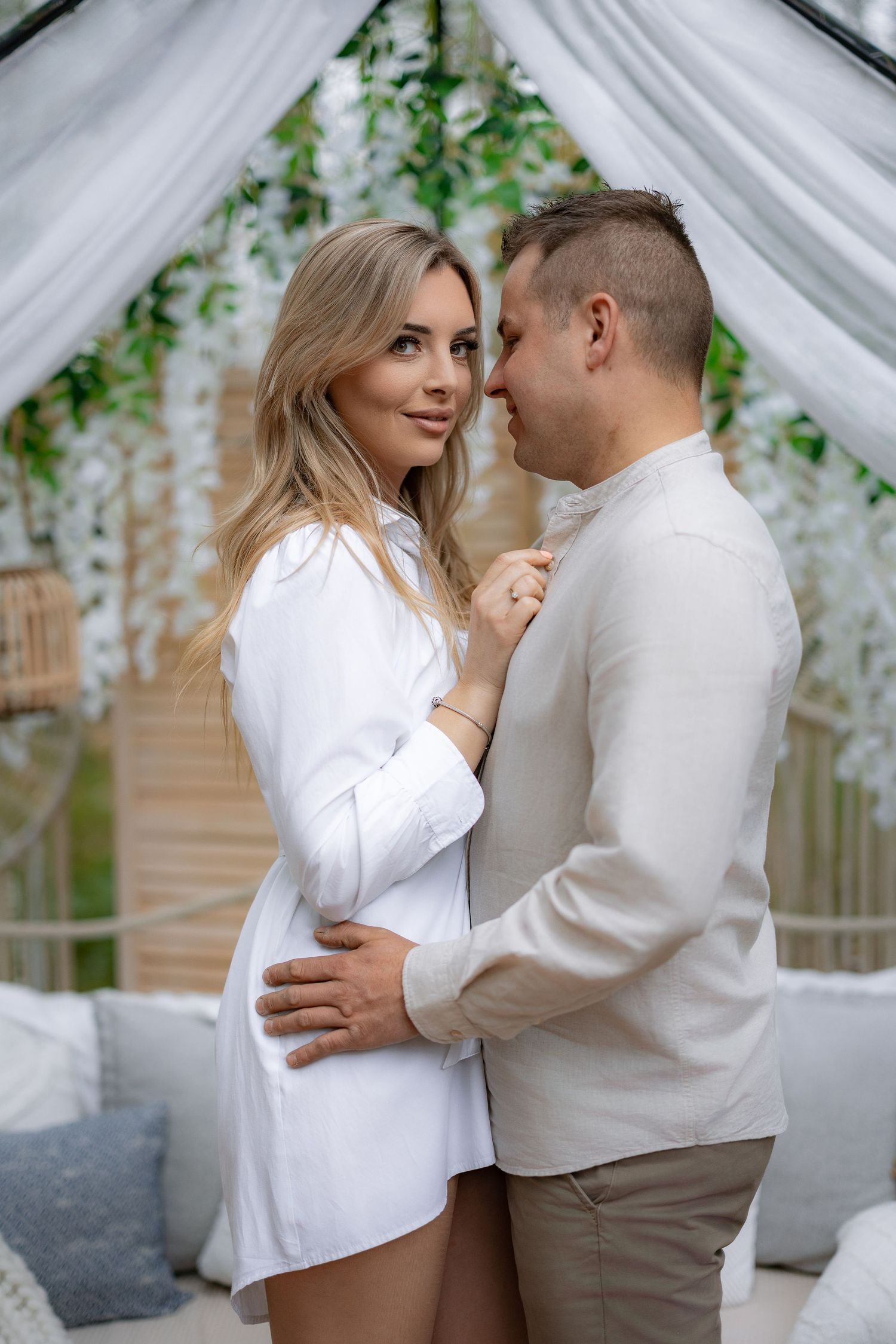 Intimate portrait of couple in white outfits under decorated wedding arch with greenery.