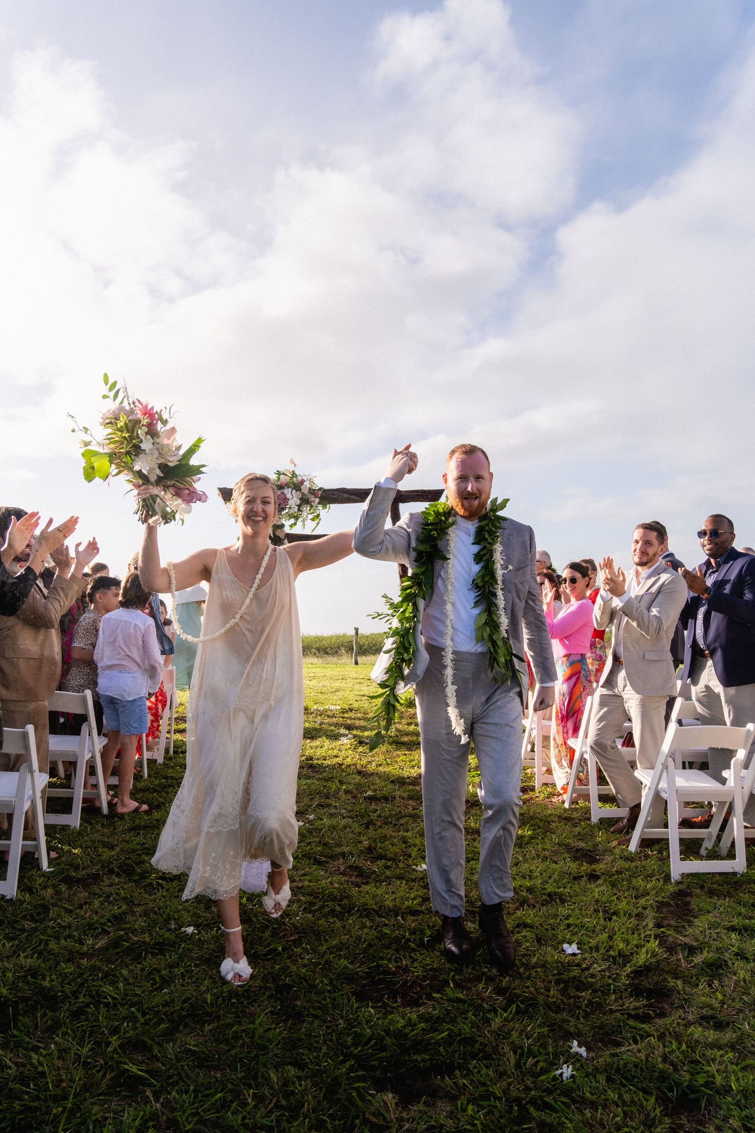 Newlyweds walk down the aisle with raised hands, wearing traditional lei garlands at an outdoor Hawaii wedding celebration.