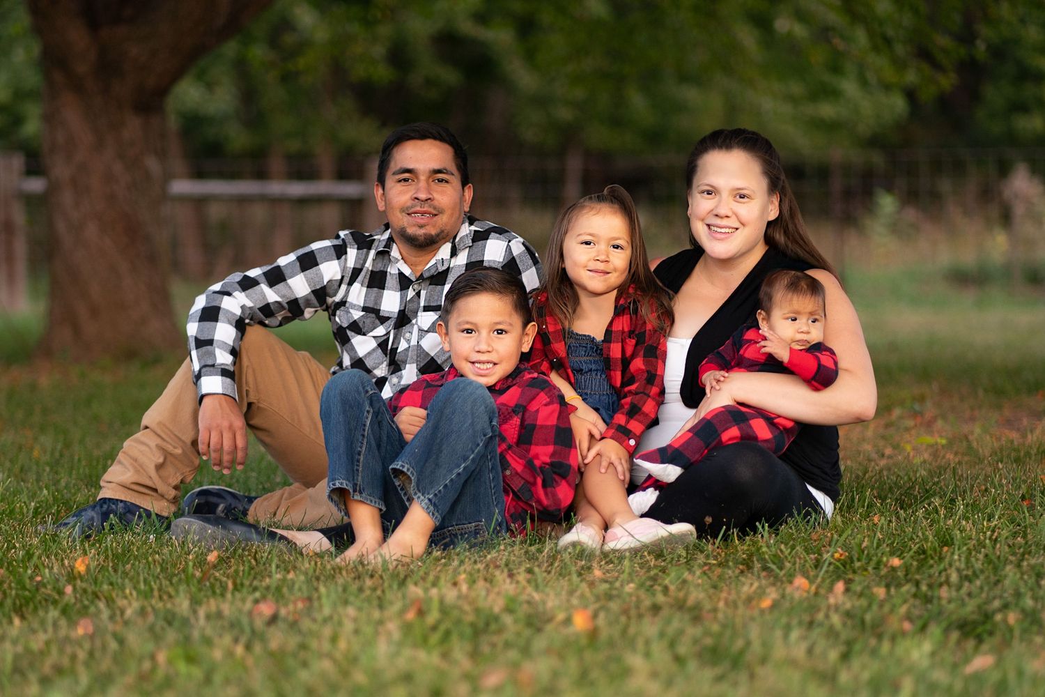 A family of five wearing coordinated red and black plaid shirts poses together on grass during an outdoor photo session.