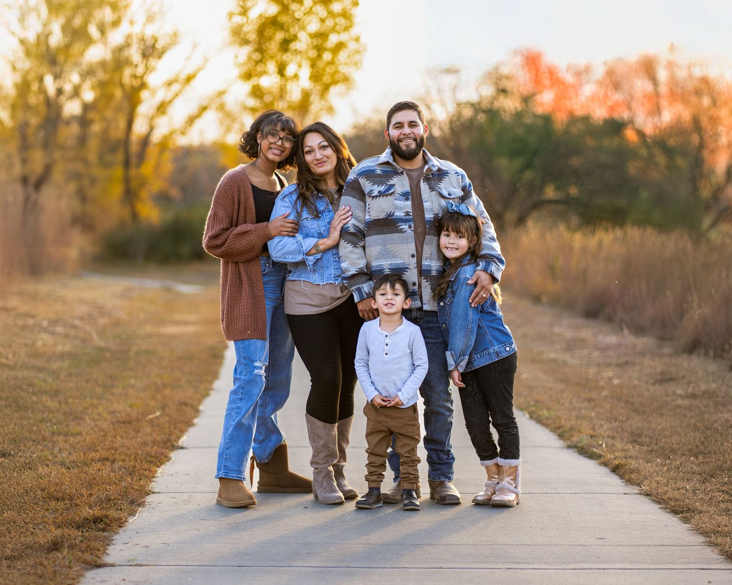 A family poses together on a paved path during sunset in autumn with golden foliage in the background.