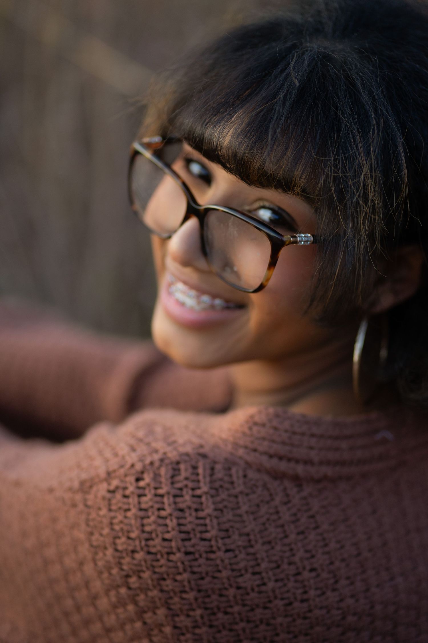Close up portrait of someone wearing glasses and a cozy brown knit sweater smiling warmly at the camera.