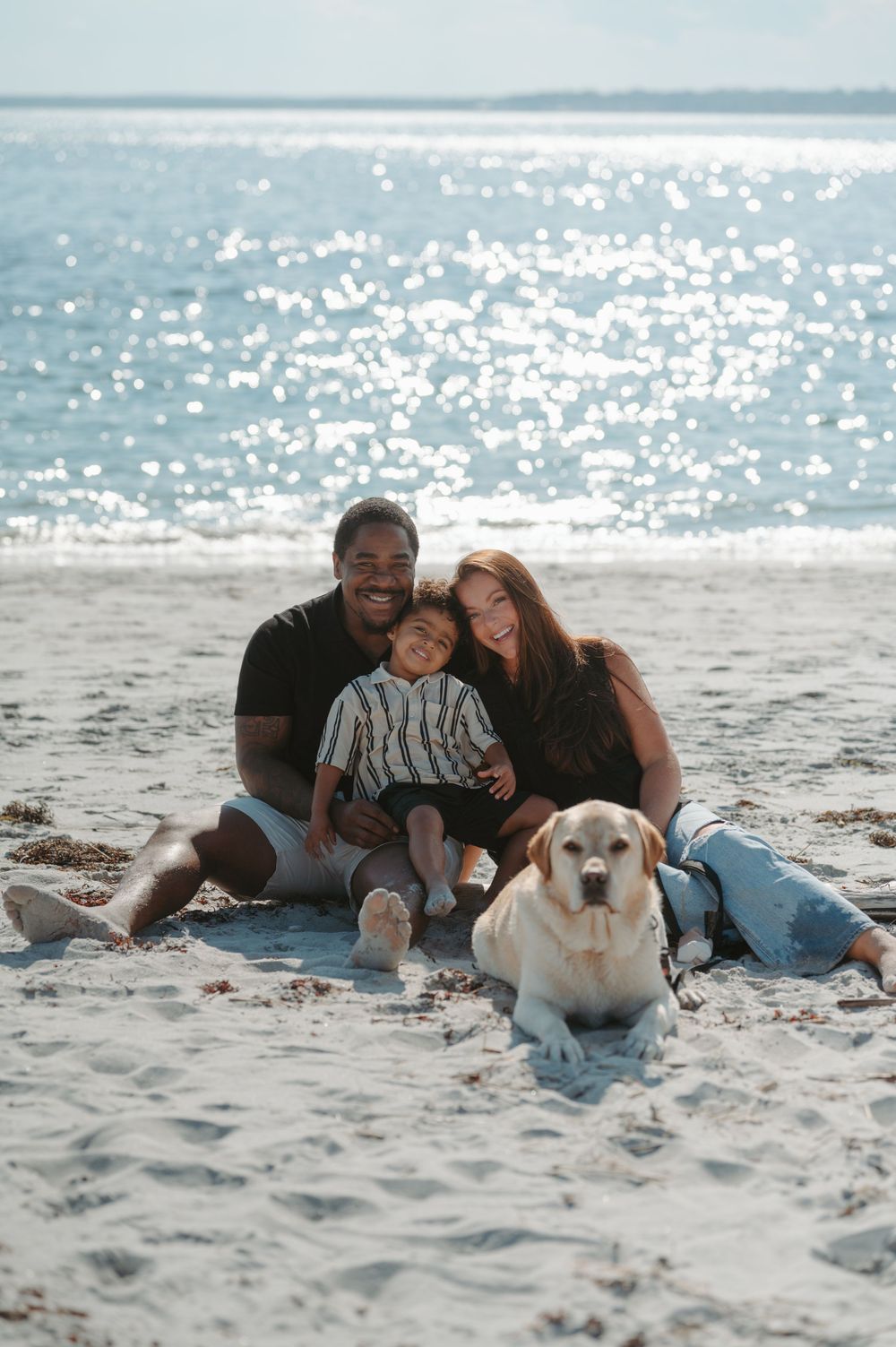 Family and their yellow Labrador dog relaxing together on a sunny beach with sparkling ocean waves in the background.