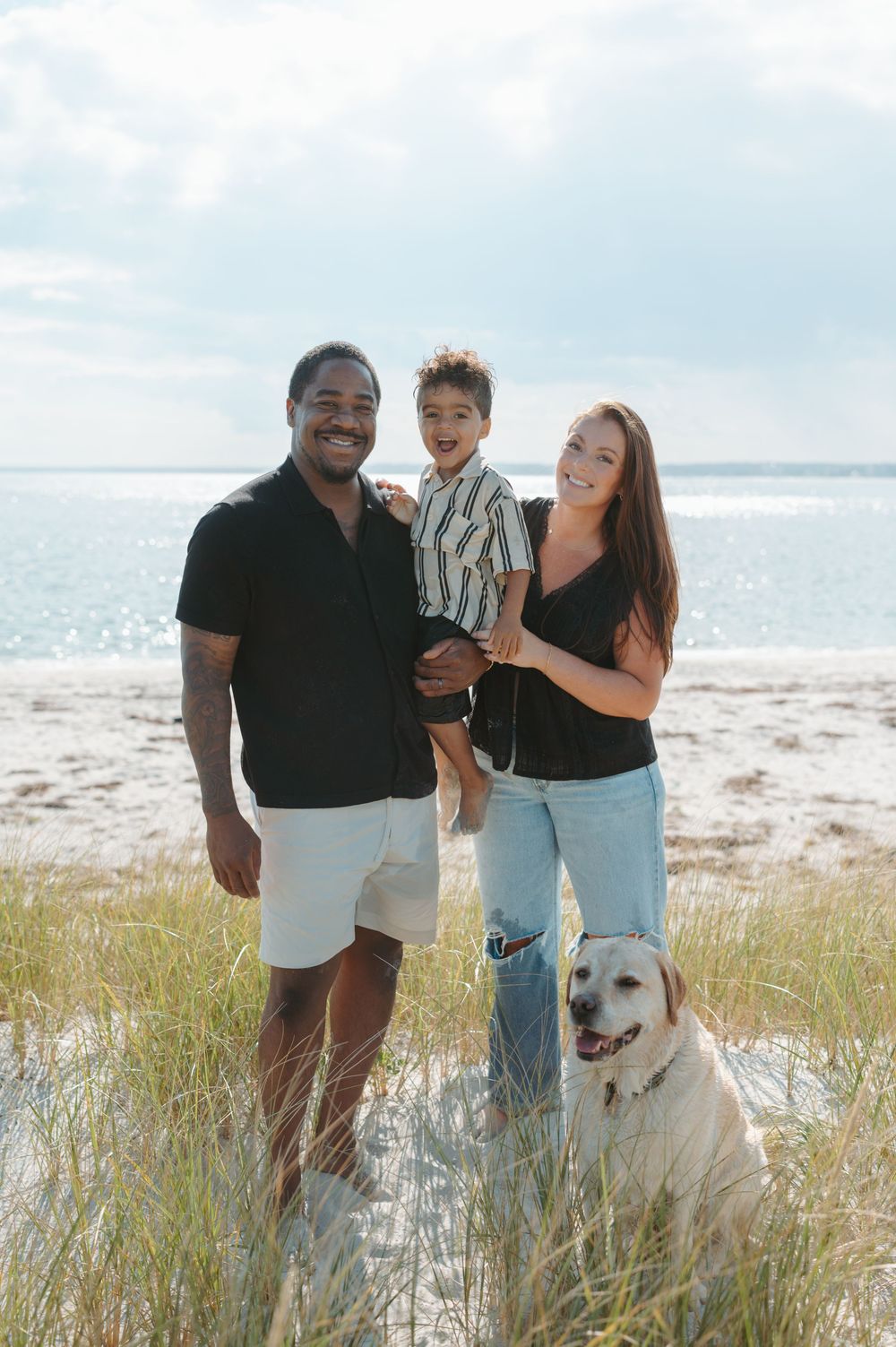 A family poses together with their white dog on a sunny beach with tall grass and sand dunes in the background.