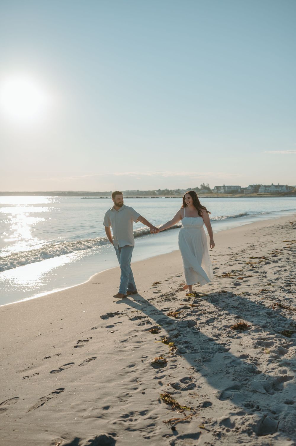 Couple walking along a sunny beach shoreline with sparkling ocean waves and clear blue sky.