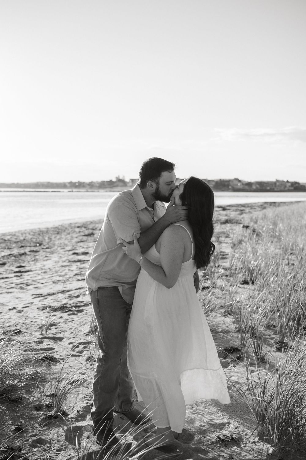 A couple embraces in a romantic moment on a scenic beach with tall grass and sand dunes in the background.