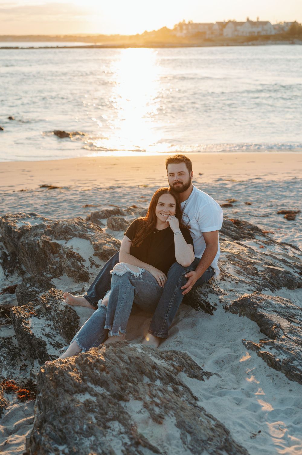 A couple sits together on rocky coastal beach at sunset enjoying golden hour light and ocean views.