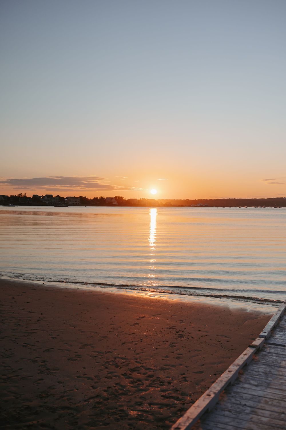 Beautiful sunset over calm ocean waters with golden light reflecting on sandy beach and wooden boardwalk.