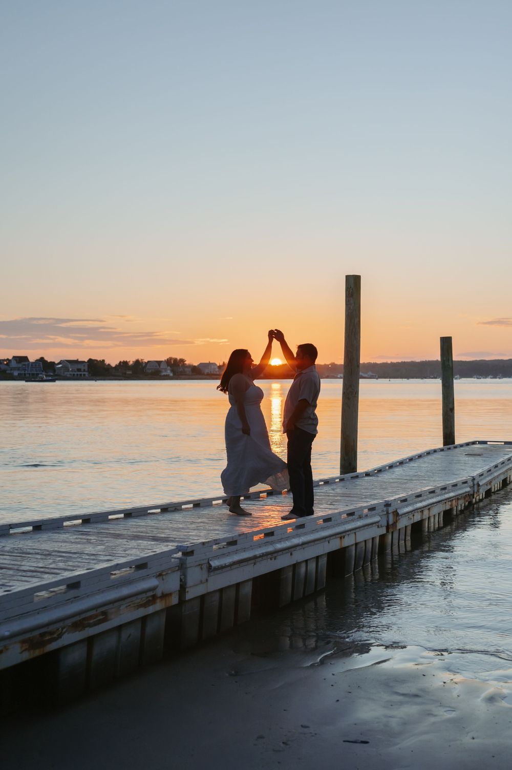 Silhouettes dance on a wooden pier at sunset with orange sky and calm waters in the background.