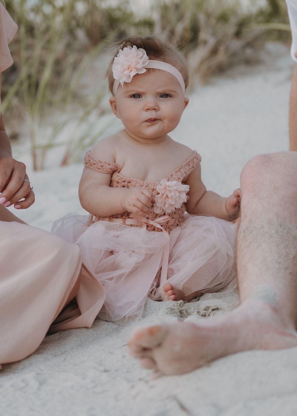 Baby in pink tulle dress with headband sits on beach sand.