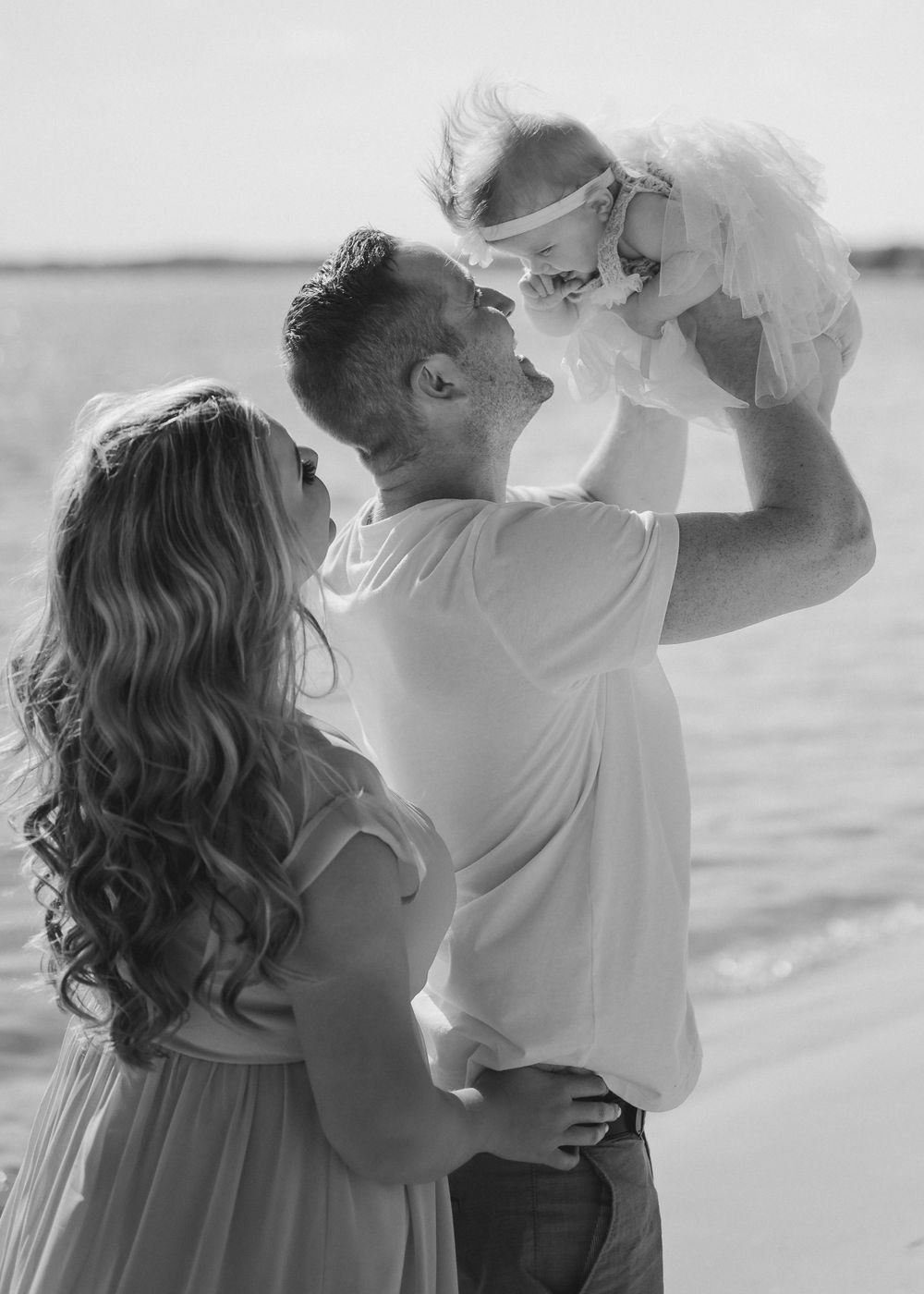 Black and white photo of family moment at beach with baby being lifted in air.