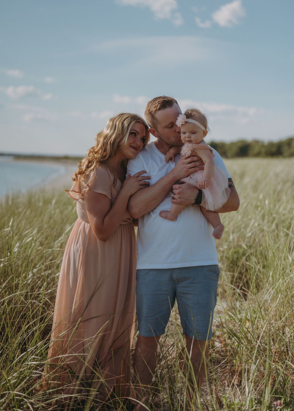 Family of three embracing in tall grass by waterfront on sunny day.