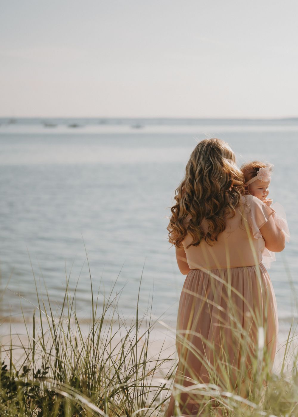 Woman in flowing dress holds baby while standing by beach at sunset.
