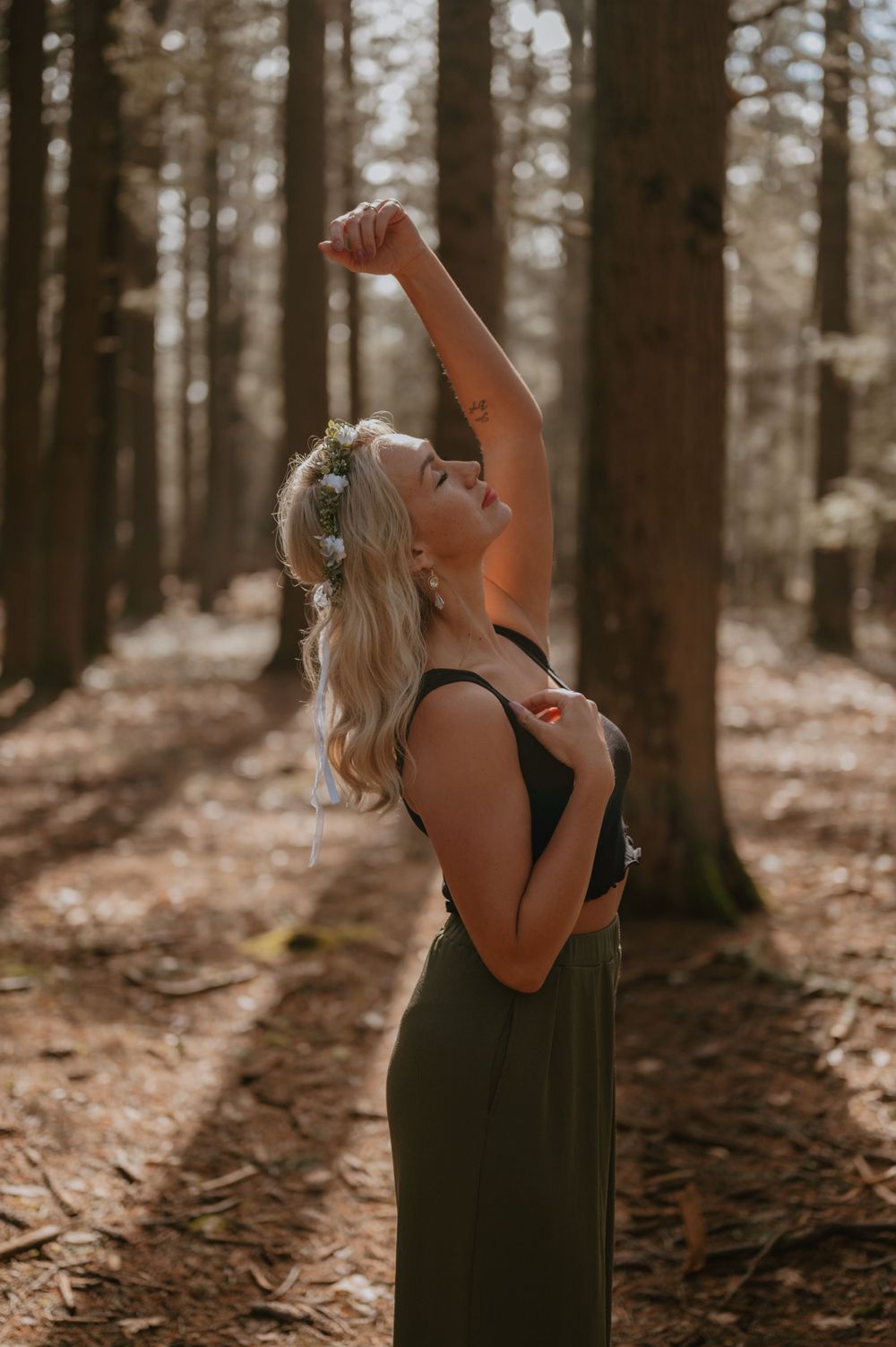 Person in forest wearing floral crown and dress reaching up towards sunlight filtering through trees.