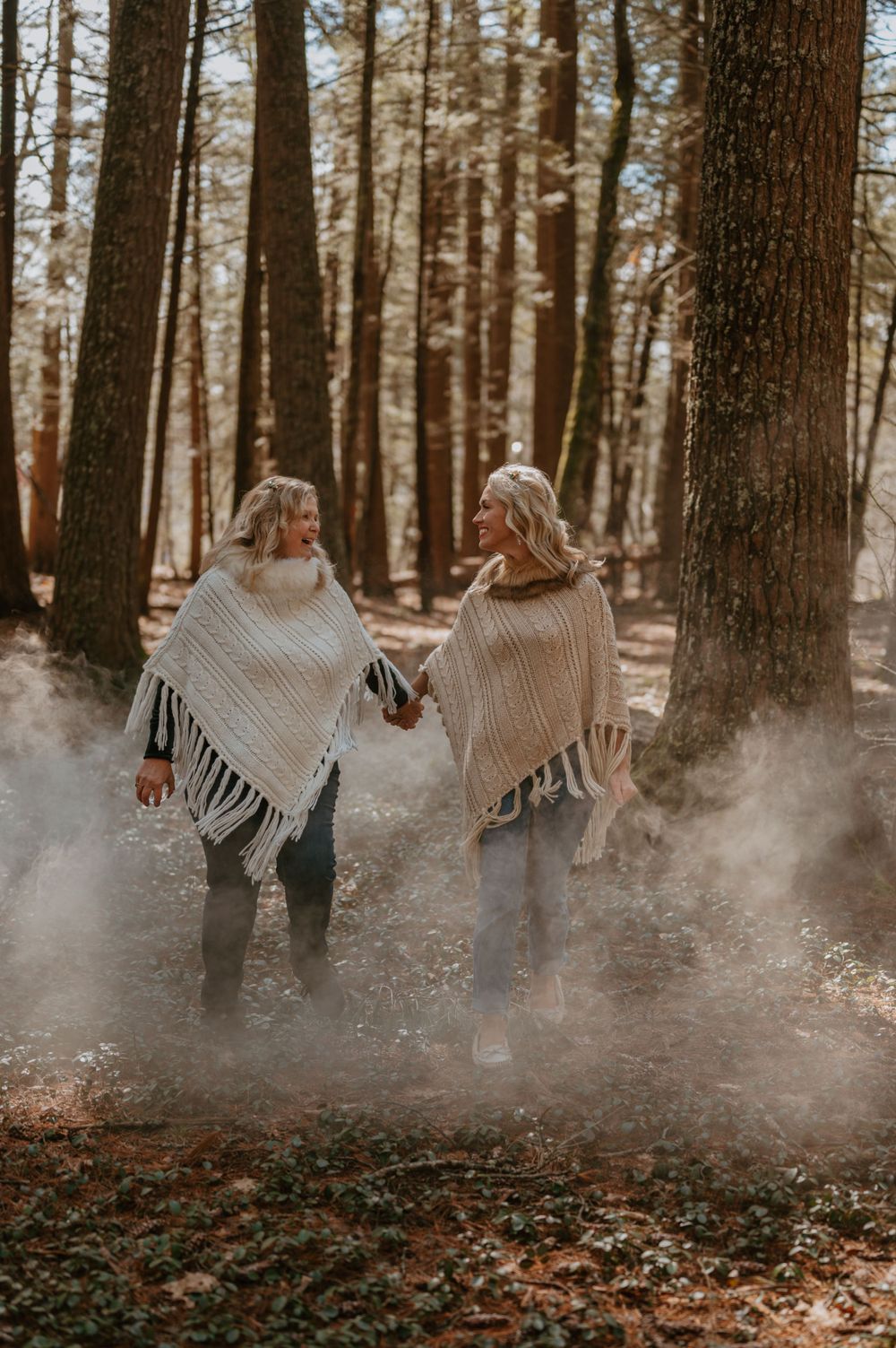 Two people in knit ponchos hold hands while walking through a misty autumn forest.