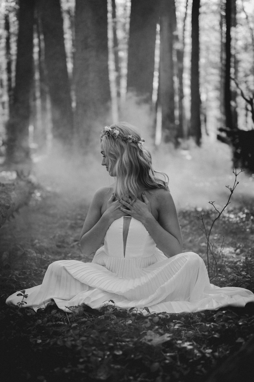 Ethereal black and white photo of a bride sitting in a misty forest wearing a white gown.