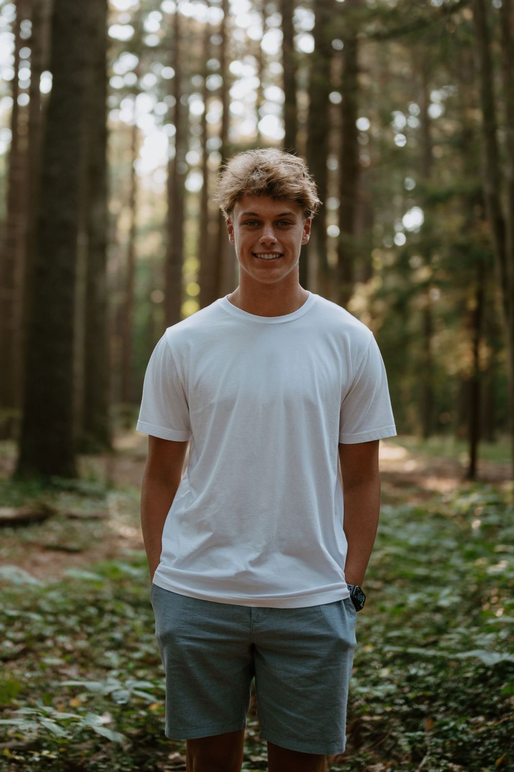 A casual outdoor portrait in a forest setting with sunlight filtering through pine trees.