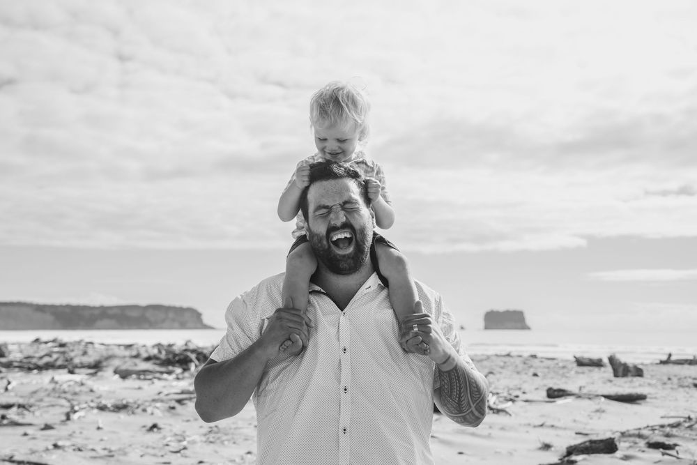 toddler sitting on dads shoulders pulling his hair and laughing at the beach