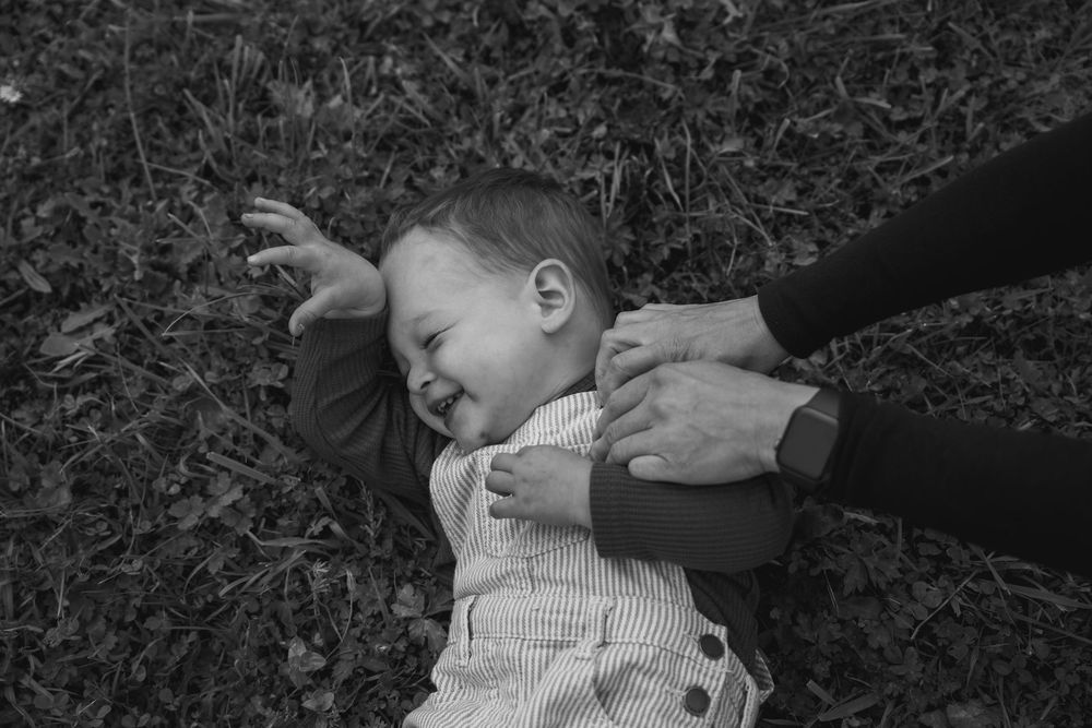 toddler laying on grass laughing with mothers hands tickling his shoulder