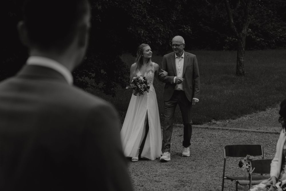 Black and white photo of bride being walked down aisle by father during outdoor ceremony.