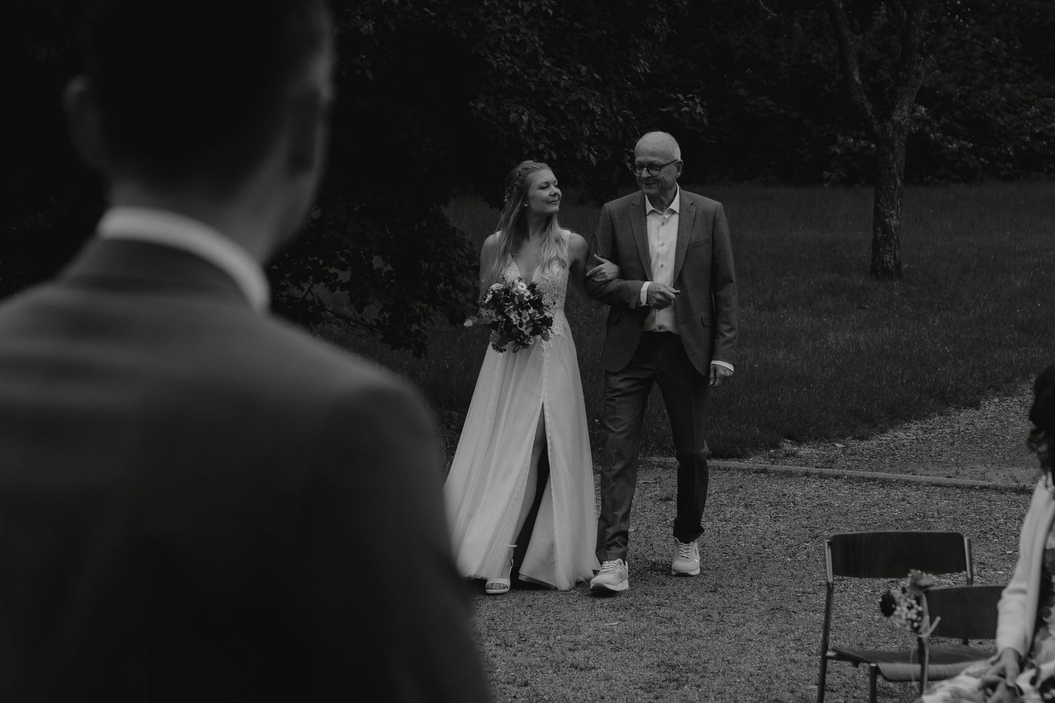 Black and white photo of bride being walked down aisle by father during outdoor ceremony.