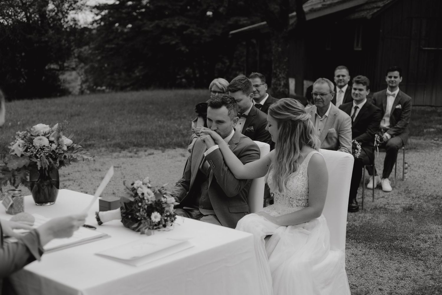 Wedding ceremony outdoors with a white-clothed table and guests gathered on a lawn.