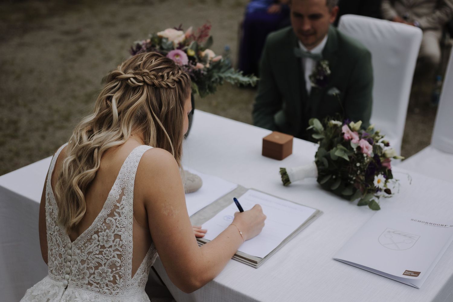Bride in lace back dress signing marriage certificate at outdoor ceremony.
