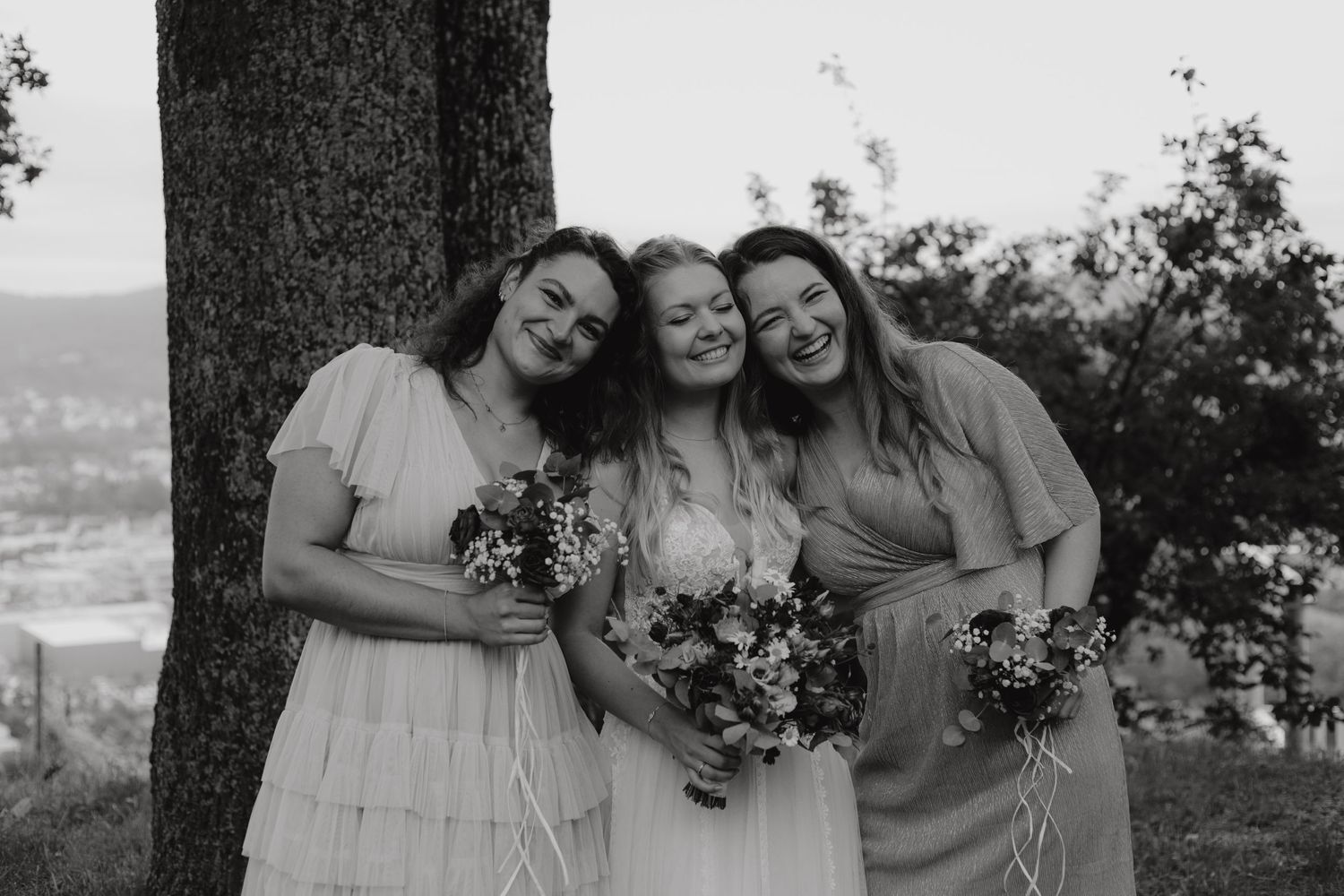 Three women in light dresses pose joyfully together outdoors with bouquets.