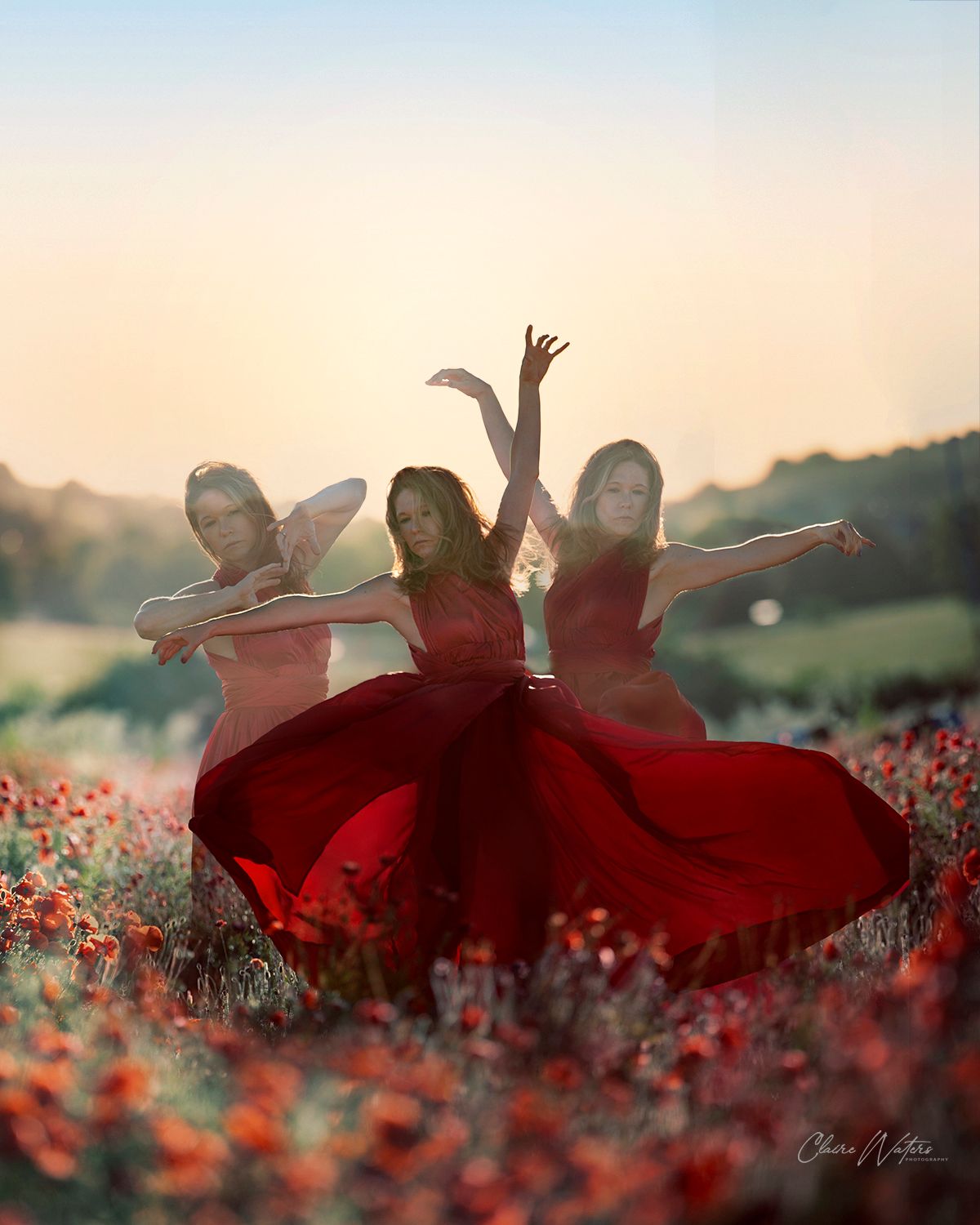 Three dancers in flowing red dresses twirl gracefully among a field of red flowers at sunset.