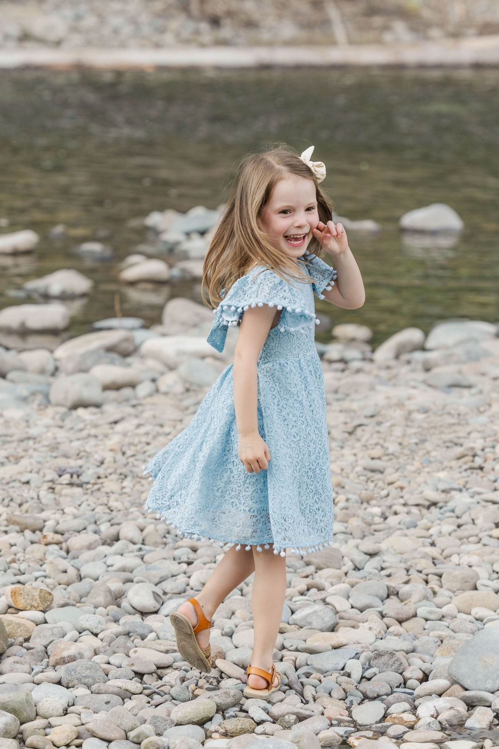 Adorable little girl in a blue lace dress smiling and walking on a rocky riverbank.