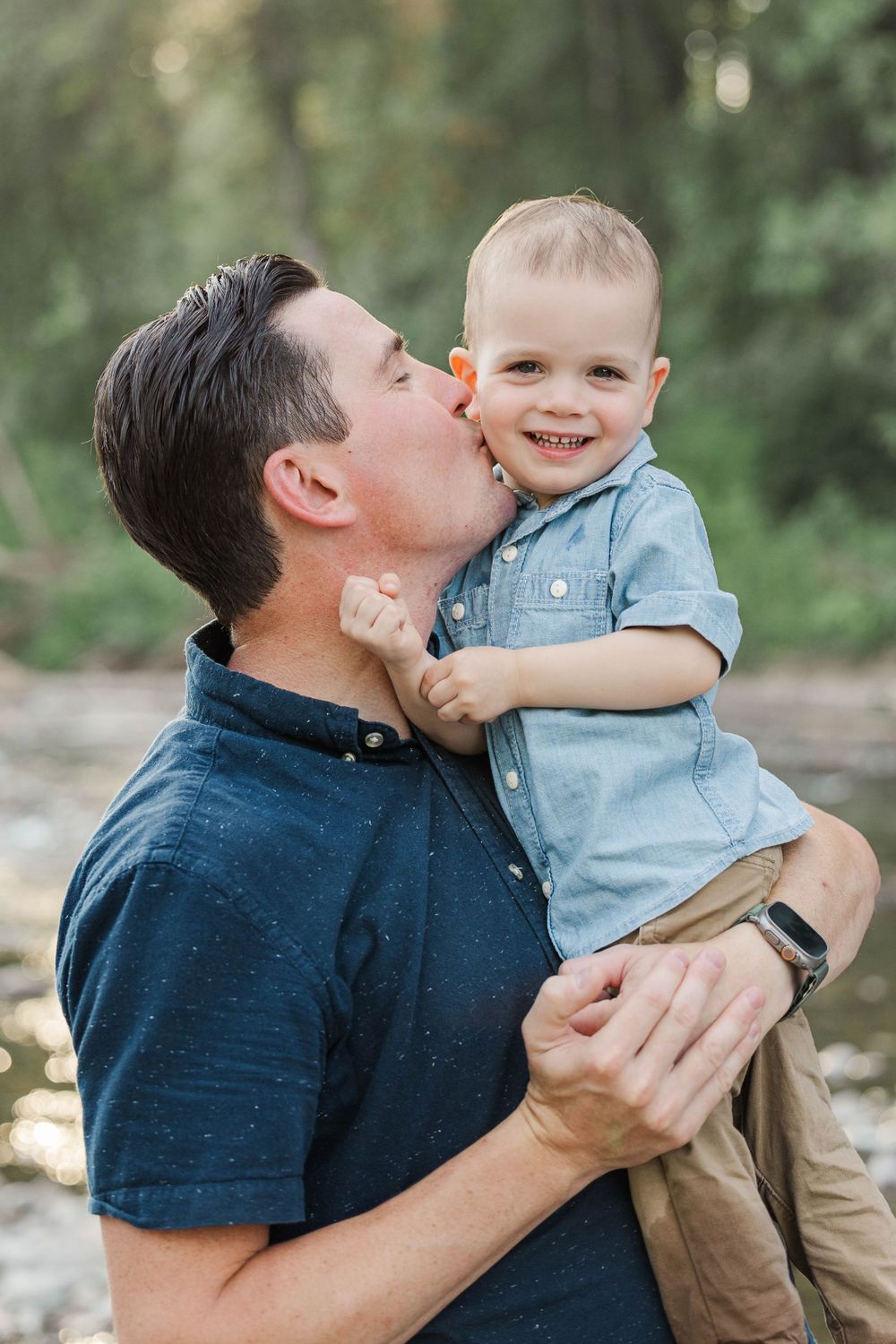 Loving father kissing smiling toddler son on the cheek while holding him outdoors near a river.