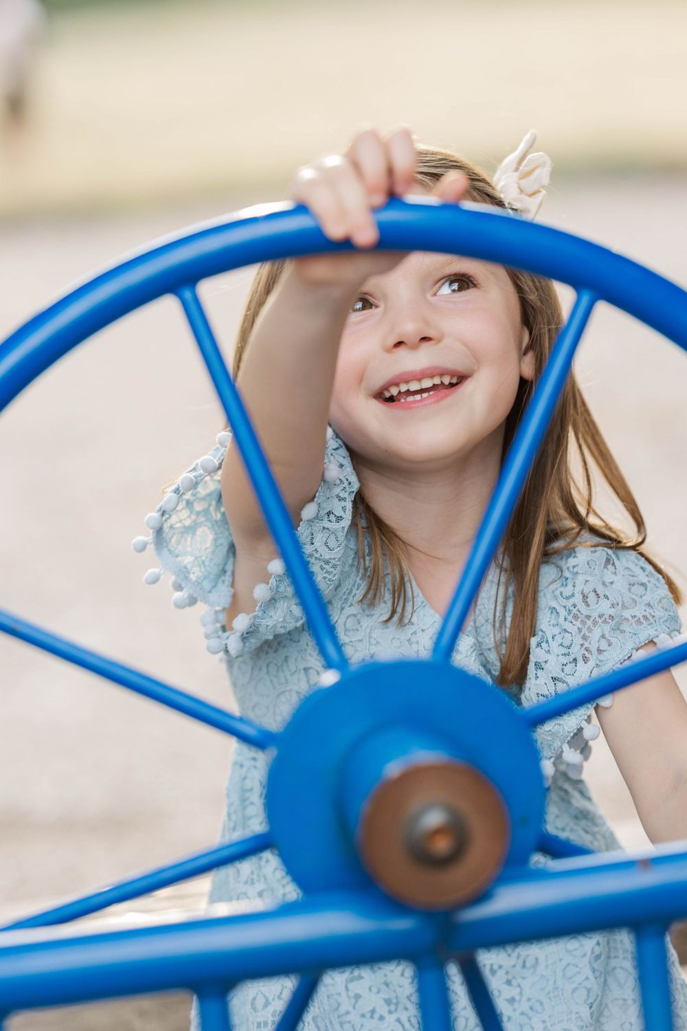 Happy young girl in a blue lace dress smiling while playing on a blue wheel at a playground.