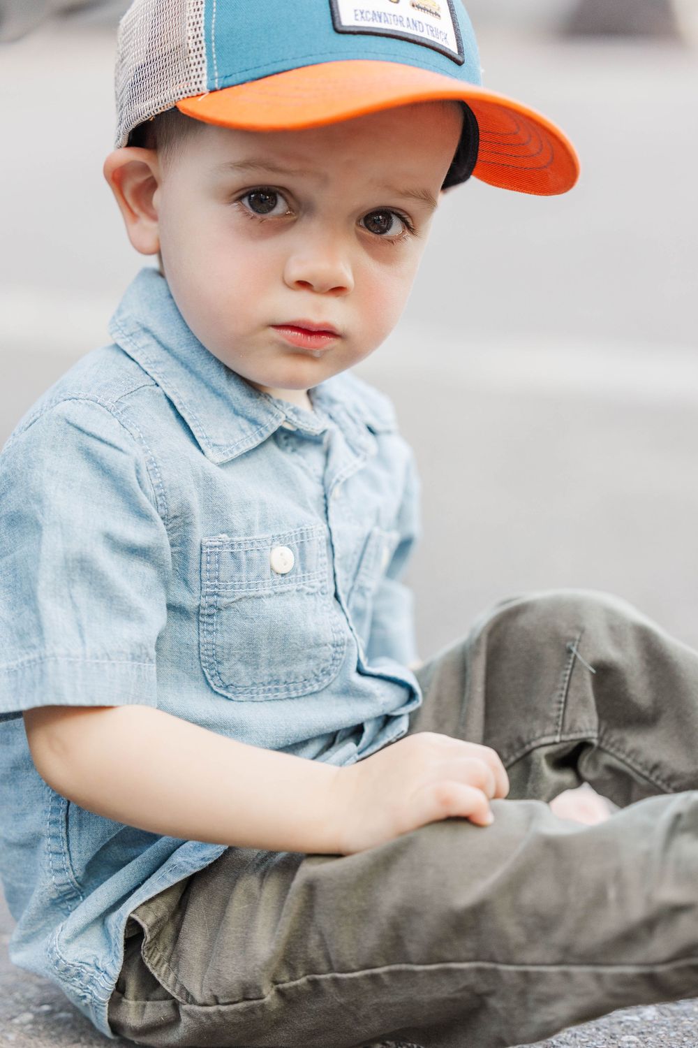 Adorable toddler boy in a blue shirt and orange cap sitting outdoors looking at the camera.