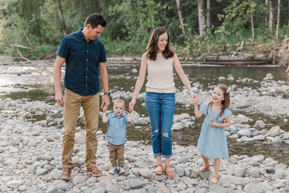 Family of four walking hand in hand along a rocky riverbank surrounded by lush greenery.