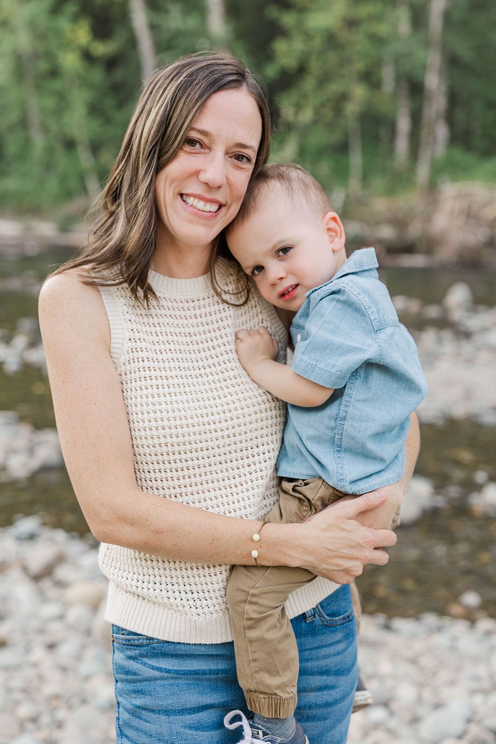 Smiling mother holding her young toddler son outdoors near a tree-lined rocky riverbank.