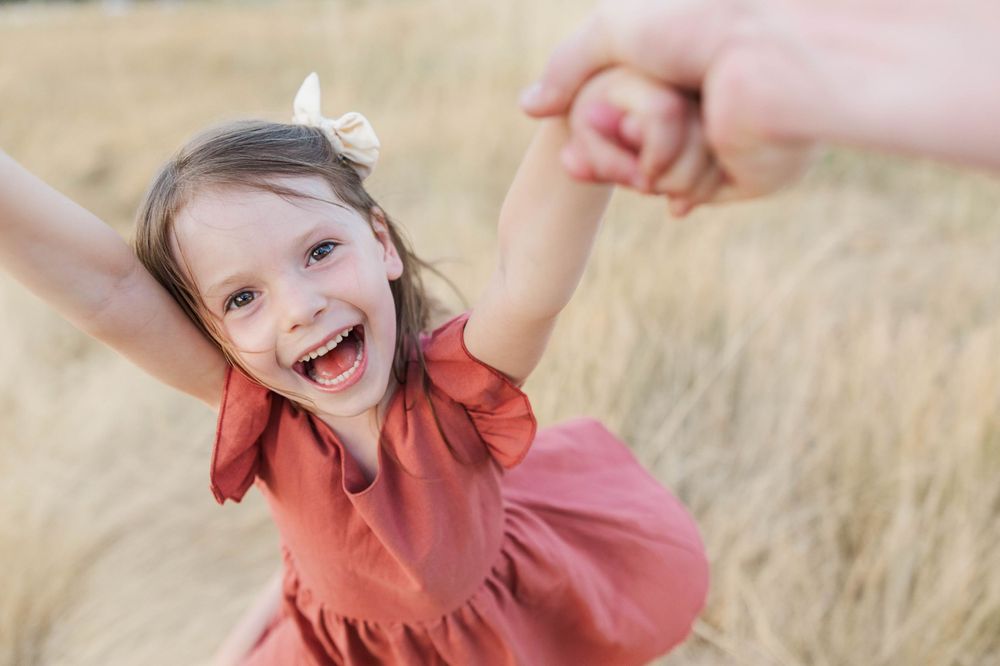Joyful little girl in a red dress laughing while being swung by her parents hands outdoors.