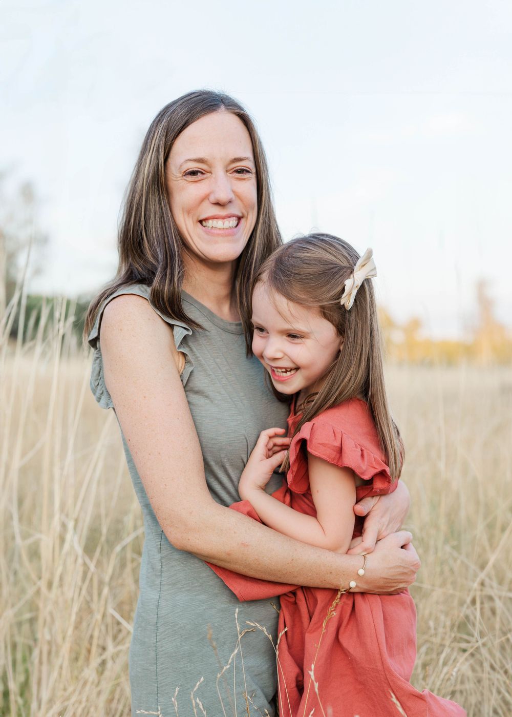 Smiling mother hugging her young daughter in a red dress in a golden grass field.