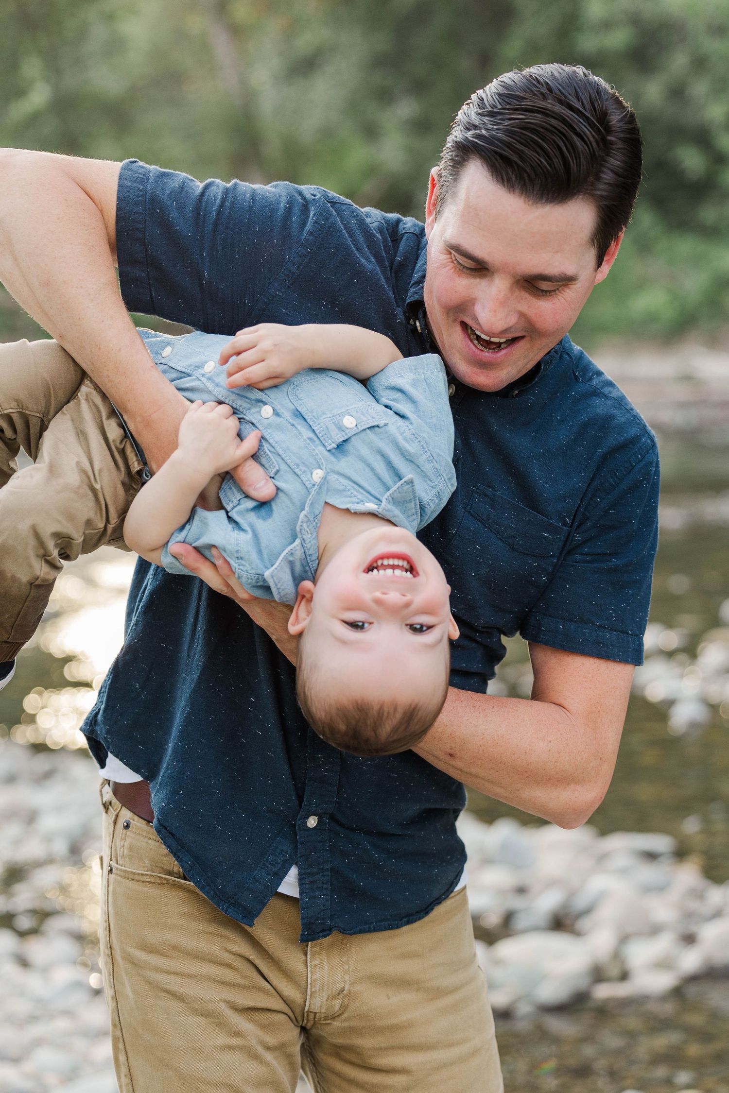 Laughing father playfully holds young son upside down by a rocky riverside outdoors.