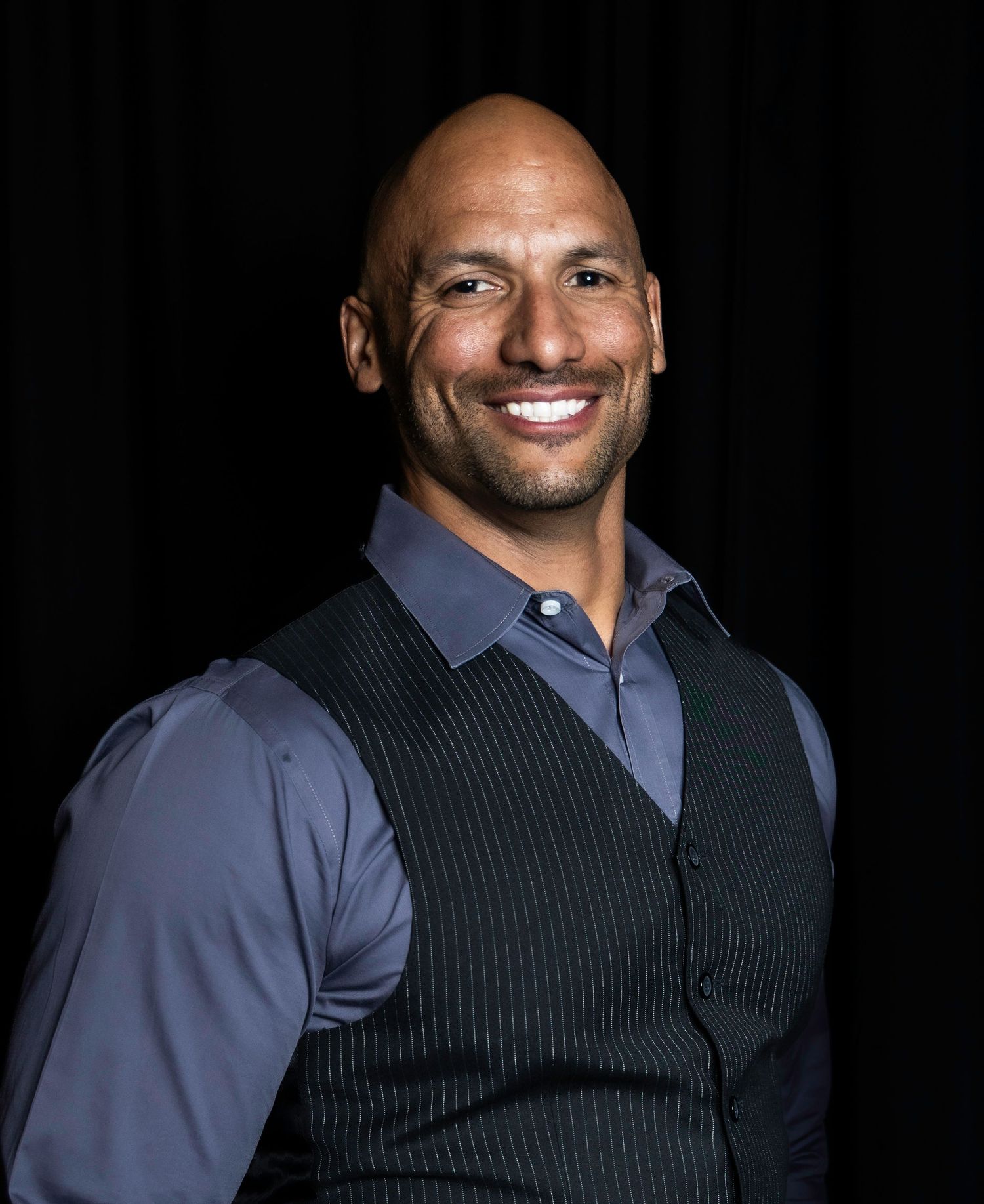 Professional portrait showing gray shirt and black vest against dark studio backdrop. Headshot photography