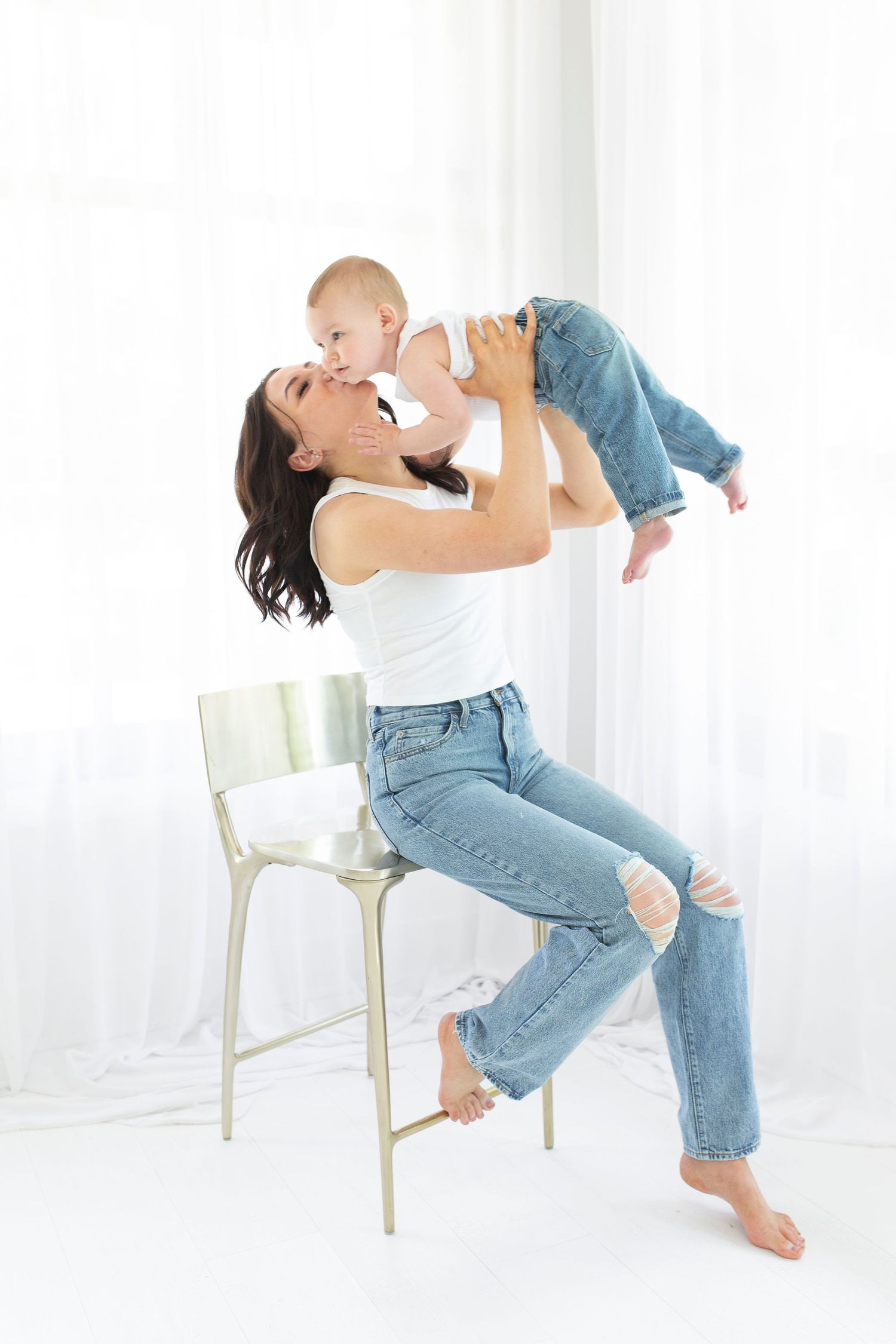 Parent playfully lifts baby in white tank top and jeans while sitting on chair against white backdrop.