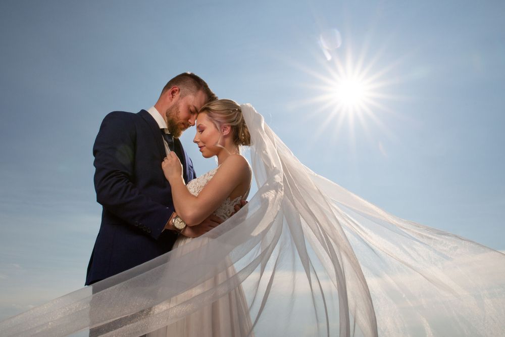 Wedding couple kisses under flowing veil with bright sunburst in blue sky.
