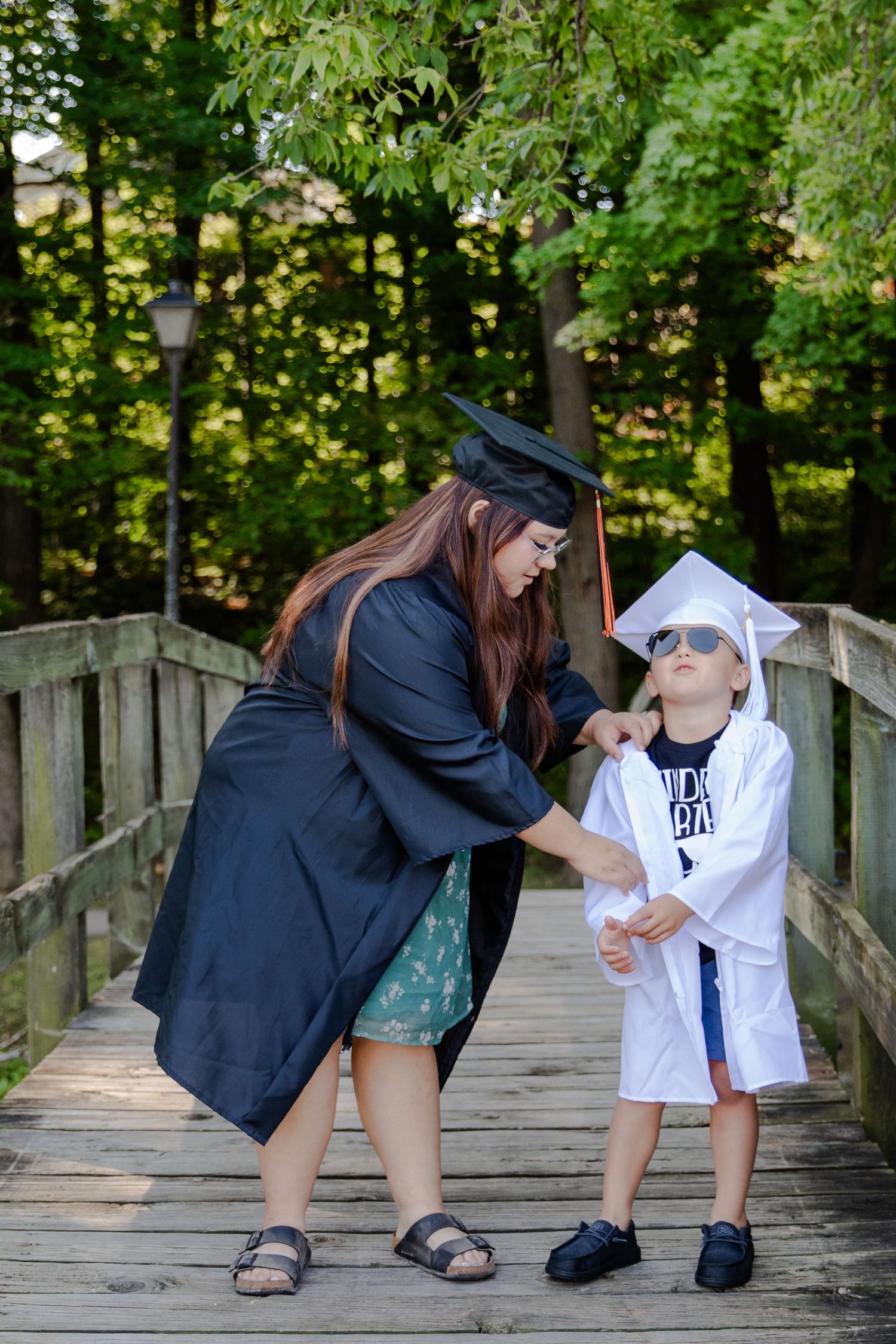 Brother and sister in their cap and gown outside on a bridge. Big sister is helping straighten little brothers gown.