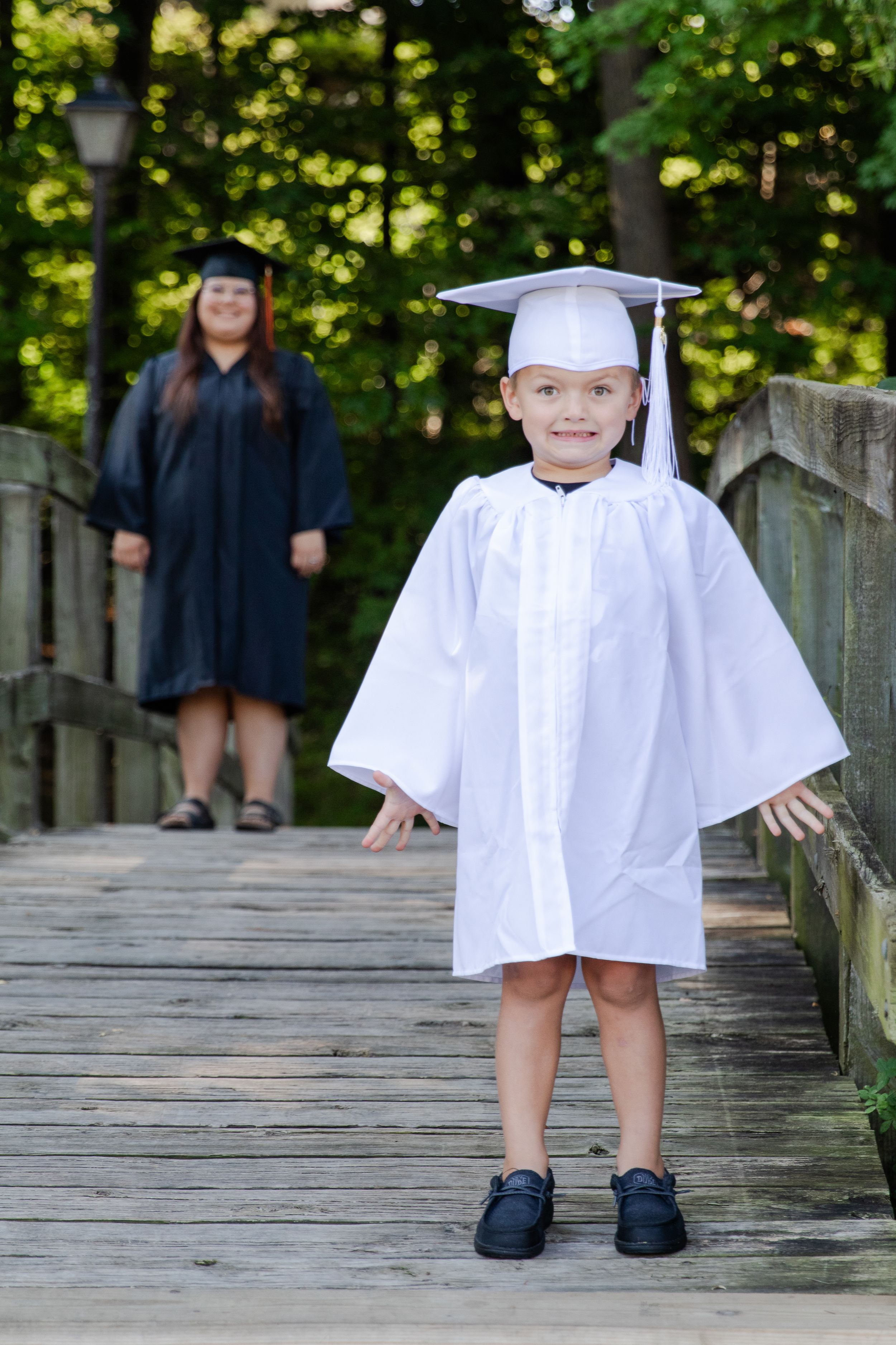 Senior and a Kindergartener in their cap and gown outside on a bridge on their first day of school.
