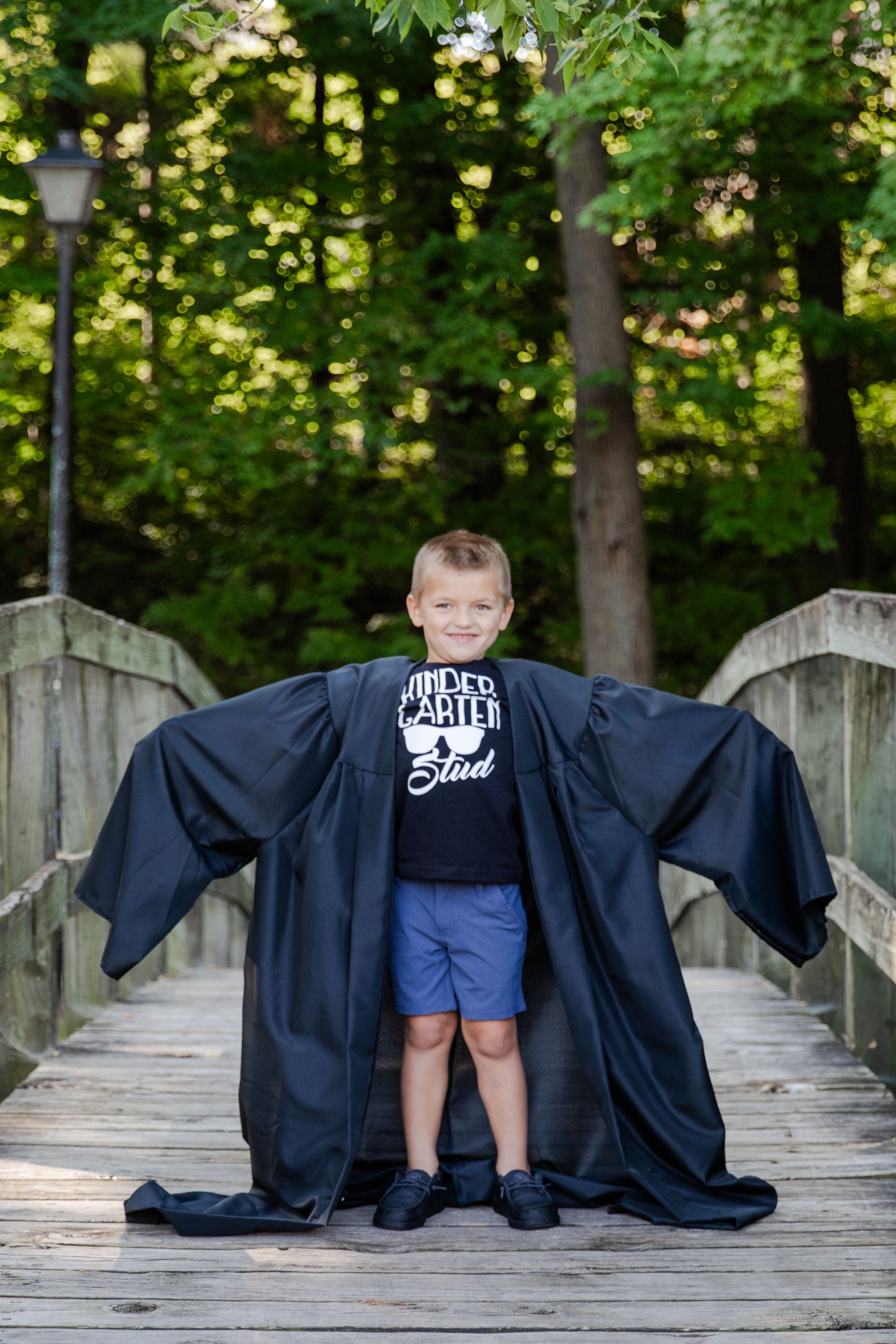 Smiling kindergartener wearing a cap and gown that's to big. The child has a lot of growing to do to fit into this gown.