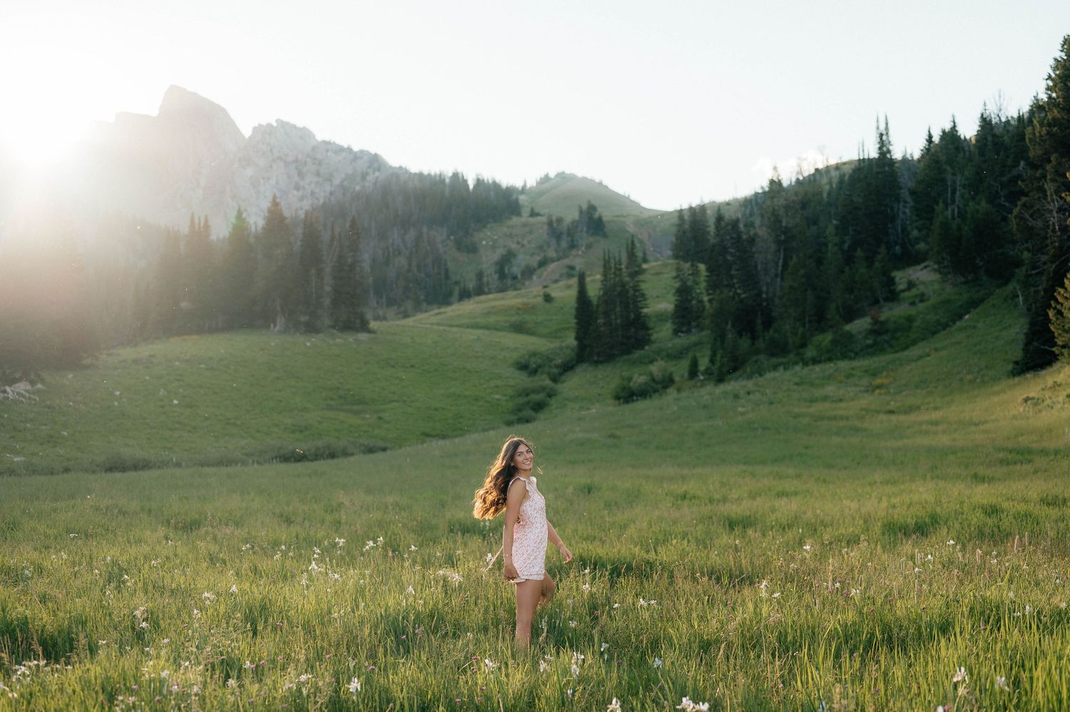 Woman in white dress standing in a wildflower meadow with dramatic mountain and pine tree scenery.